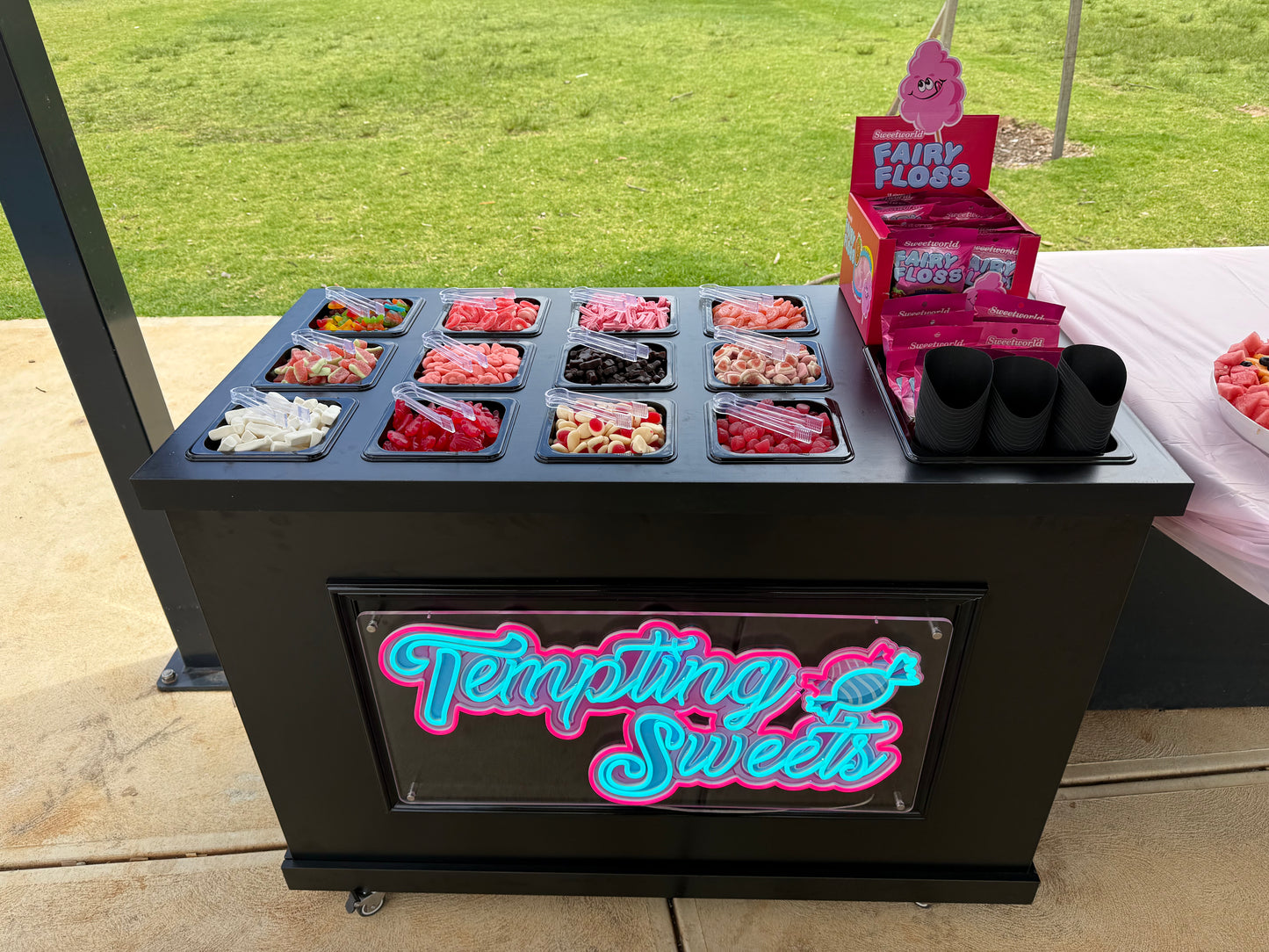 A black Tempting Sweets candy cart with a pink and blue neon-style sign on the front, filled with assorted candies in trays. Pink boxes and a display stand labeled Fair Floss are placed on top, with an outdoor grassy background visible behind the cart.