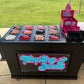 A black Tempting Sweets candy cart with a pink and blue neon-style sign on the front, filled with assorted candies in trays. Pink boxes and a display stand labeled Fair Floss are placed on top, with an outdoor grassy background visible behind the cart.