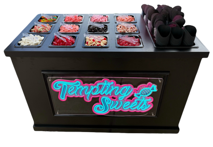 A black candy and sweets station display case with a neon-style 'Tempting Sweets' sign on the front. The top surface holds multiple clear plastic bins filled with assorted colorful candies, sweets, and pink cone stands, photographed on a plain white background.