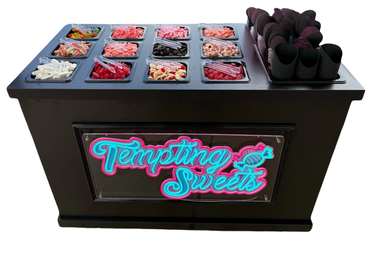 A black candy and sweets station display case with a neon-style 'Tempting Sweets' sign on the front. The top surface holds multiple clear plastic bins filled with assorted colorful candies, sweets, and pink cone stands, photographed on a plain white background.